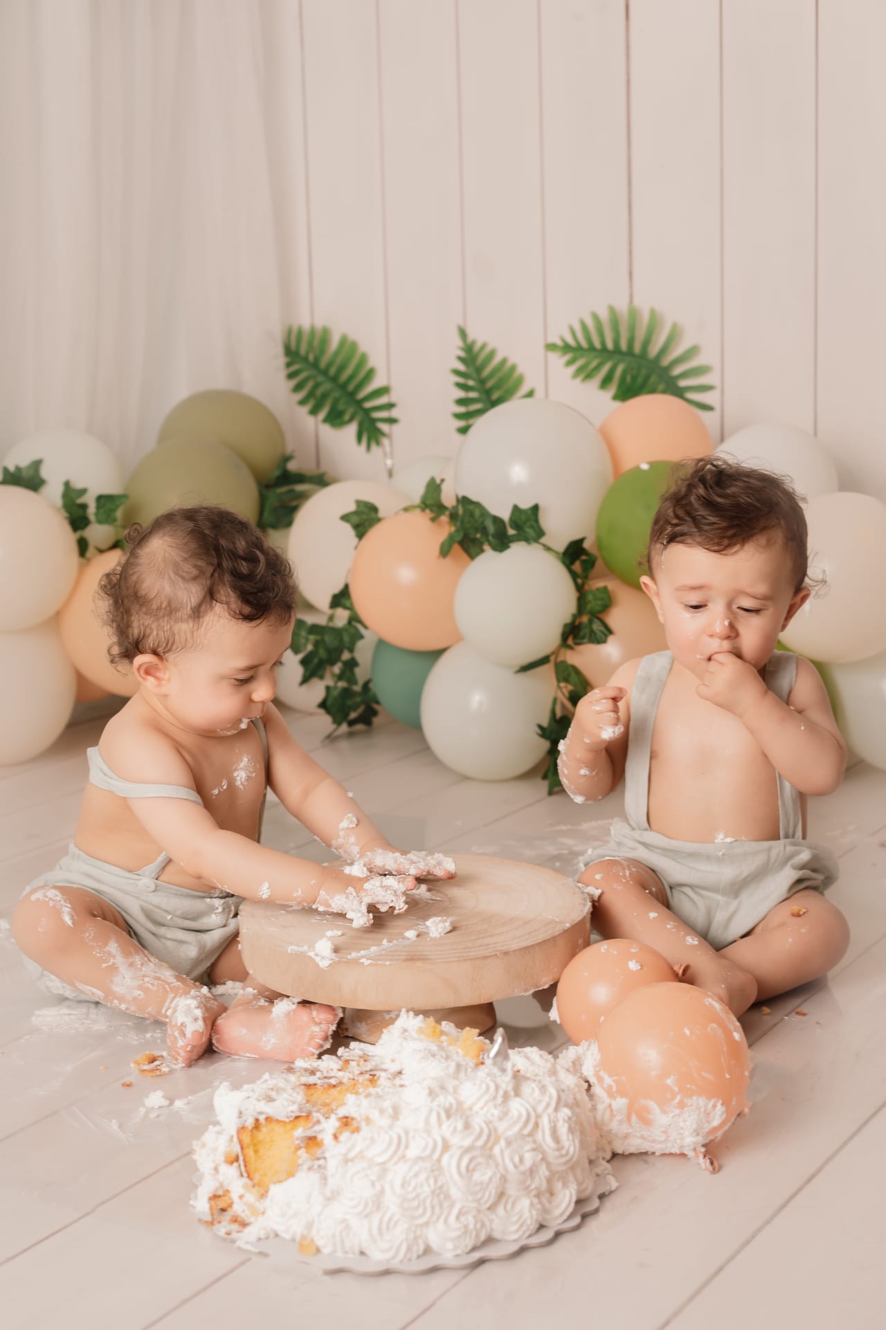 Bebés gémeos com jardineiras verde-musgo a esmagar bolo branco em base de madeira, balões pastel e folhagem ao fundo, sessão smash the cake em estúdio, Susana Schmitz Fotografia, Porto