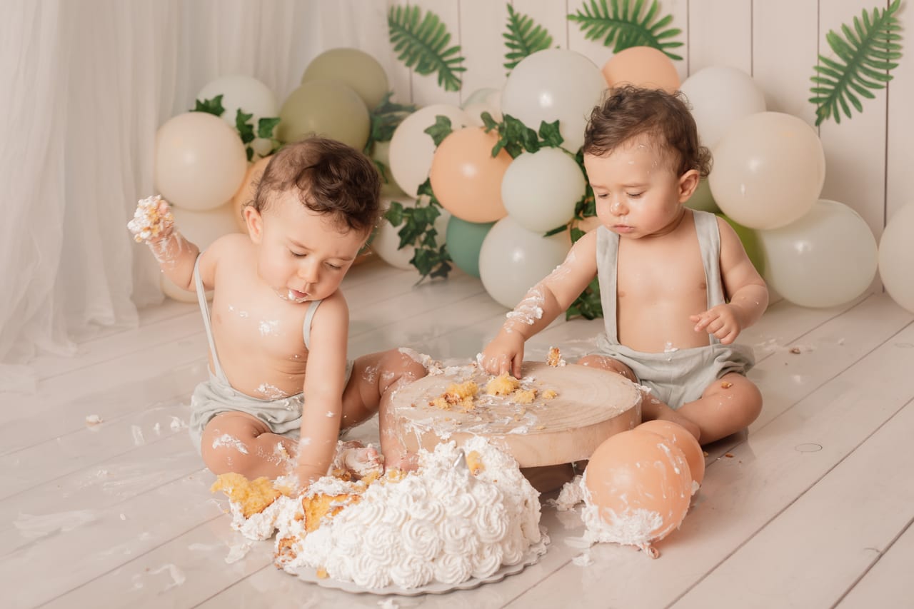 Bebés gémeos com jardineiras verde-musgo cheios de bolo a brincar com fatias caídas no chão, balões pastel e folhagem ao fundo, sessão smash the cake, Susana Schmitz Fotografia, Porto