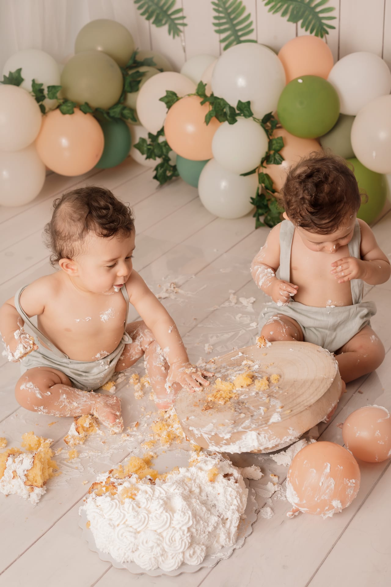 Vista de cima de bebés gémeos com jardineiras verde-musgo a desfazer bolo branco em base de madeira sobre chão branco com migalhas, sessão smash the cake, Susana Schmitz Fotografia, Porto