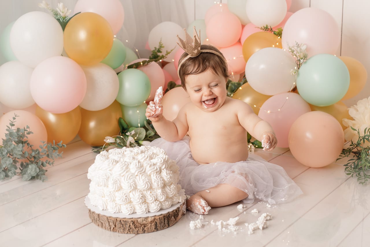 Bebé com coroa dourada e tutu branco junto a bolo branco em base de madeira, arco de balões pastel e luzes de fadas, sessão smash the cake do primeiro aniversário, Susana Schmitz Fotografia, Porto