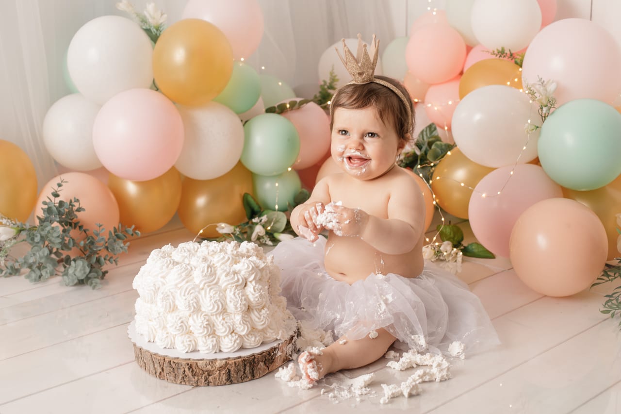 Bebé sorridente com coroa dourada e tutu branco a saborear cobertura do bolo, balões pastel e luzes de fadas em estúdio, sessão smash the cake do primeiro aniversário, Susana Schmitz Fotografia, Porto