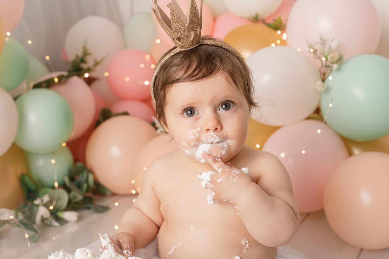 Close-up de bebé com coroa dourada com cara cheia de cobertura branca a chupar o dedo, balões pastel e luzes de fadas ao fundo, sessão smash the cake, Susana Schmitz Fotografia, Porto