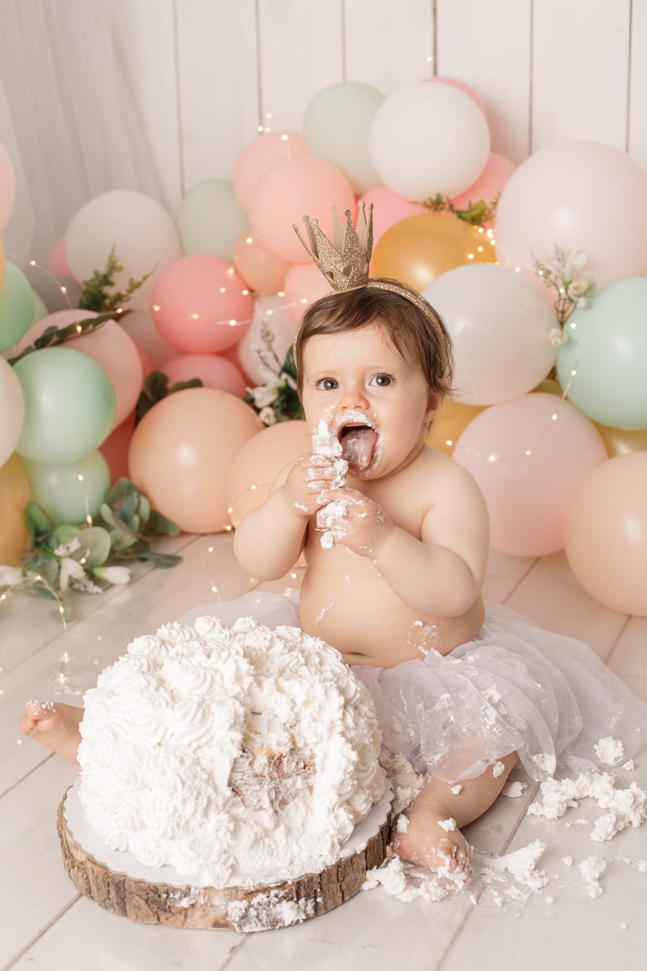 Bebé com coroa dourada e tutu branco a meter as mãos cheias de cobertura na boca após esmagar o bolo, balões pastel e luzes de fadas, sessão smash the cake, Susana Schmitz Fotografia, Porto