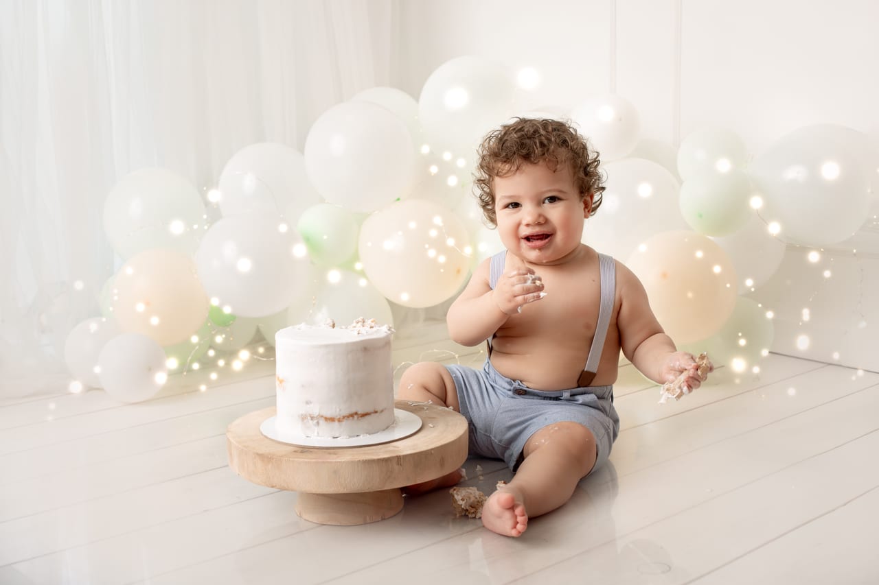 Bebé com coroa dourada e saia de tule branca junto a bolo branco sobre base de madeira, arco de balões pastel e luzes de fadas, sessão smash the cake do primeiro aniversário, Susana Schmitz Fotografia, Porto