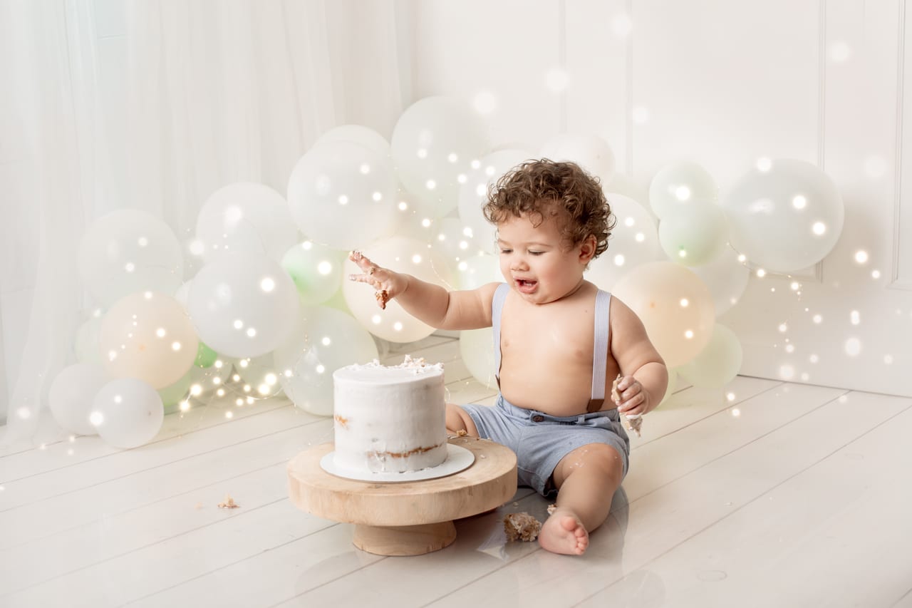 Bebé encaracolado com calções azul-claro com suspensórios sentado a olhar para bolo branco intacto em base de madeira, balões brancos com luzes de fadas, sessão smash the cake, Susana Schmitz Fotografia