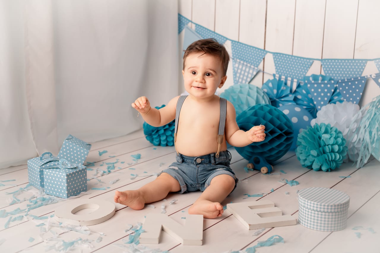 Bebé com suspensórios e calções de ganga entre bandeirolas e pompons azul-pastel com letras ONE e caixa de prendas, cenário de primeiro aniversário em estúdio, Susana Schmitz Fotografia