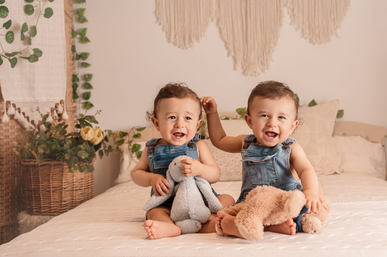 Gémeos sorridentes em jardineiras de ganga sobre cama com tapeçaria de macramé e plantas, abraçados a peluches de coelho, sessão de bebé em estúdio, Susana Schmitz Fotografia, Matosinhos