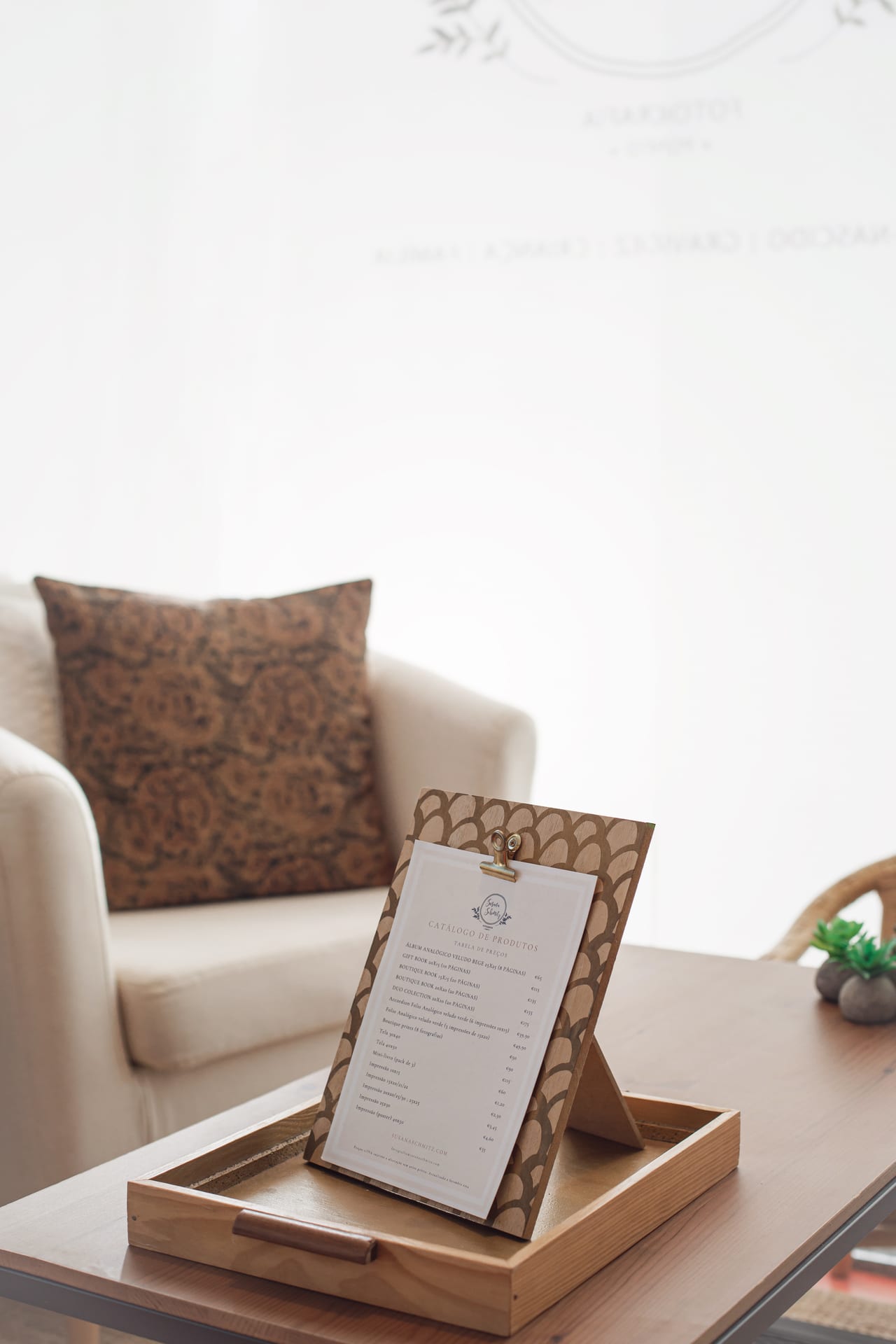 Catálogo de produtos e tabela de preços apresentado num tray de madeira na sala de receção, ao lado do cadeirão creme com almofada estampada, estúdio Susana Schmitz, Porto