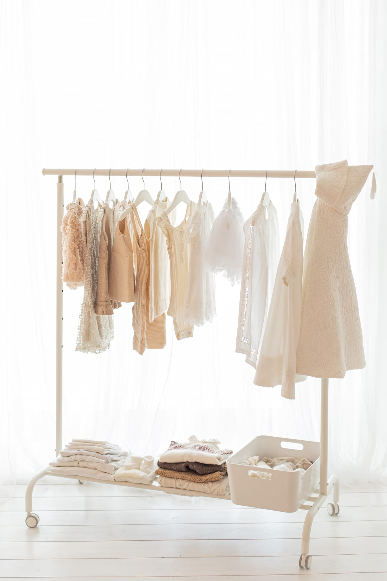 Rack de guarda-roupa para bebé e criança com peças em renda, linho e malha em tons cremes e beges, contra cortinado branco do estúdio Susana Schmitz, Leça da Palmeira