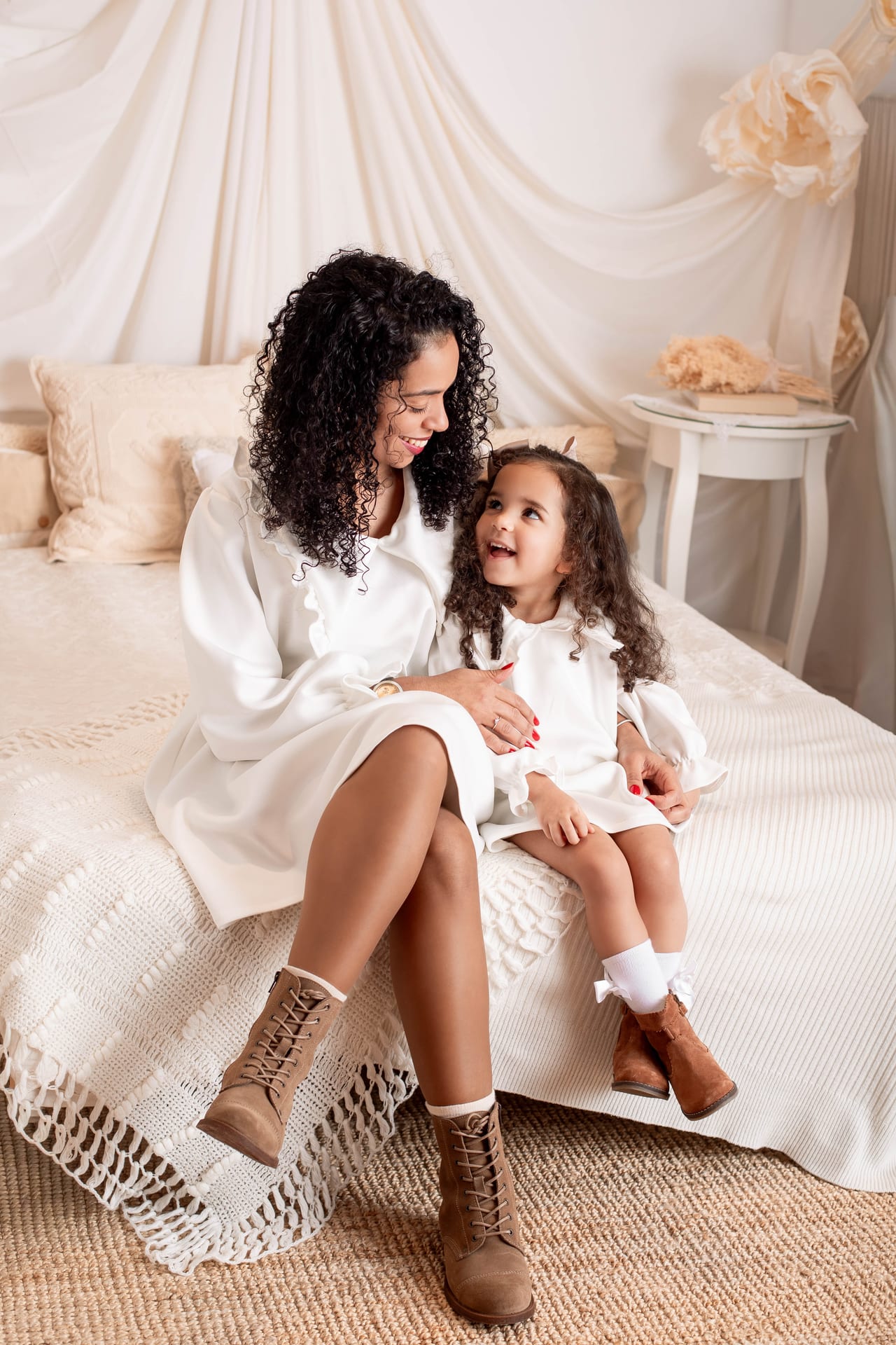 Mãe encaracolada e filha pequena em vestidos brancos com botas castanhas sobre cama com cortinas drapeadas, sessão de família em estúdio, Susana Schmitz Fotografia, Porto