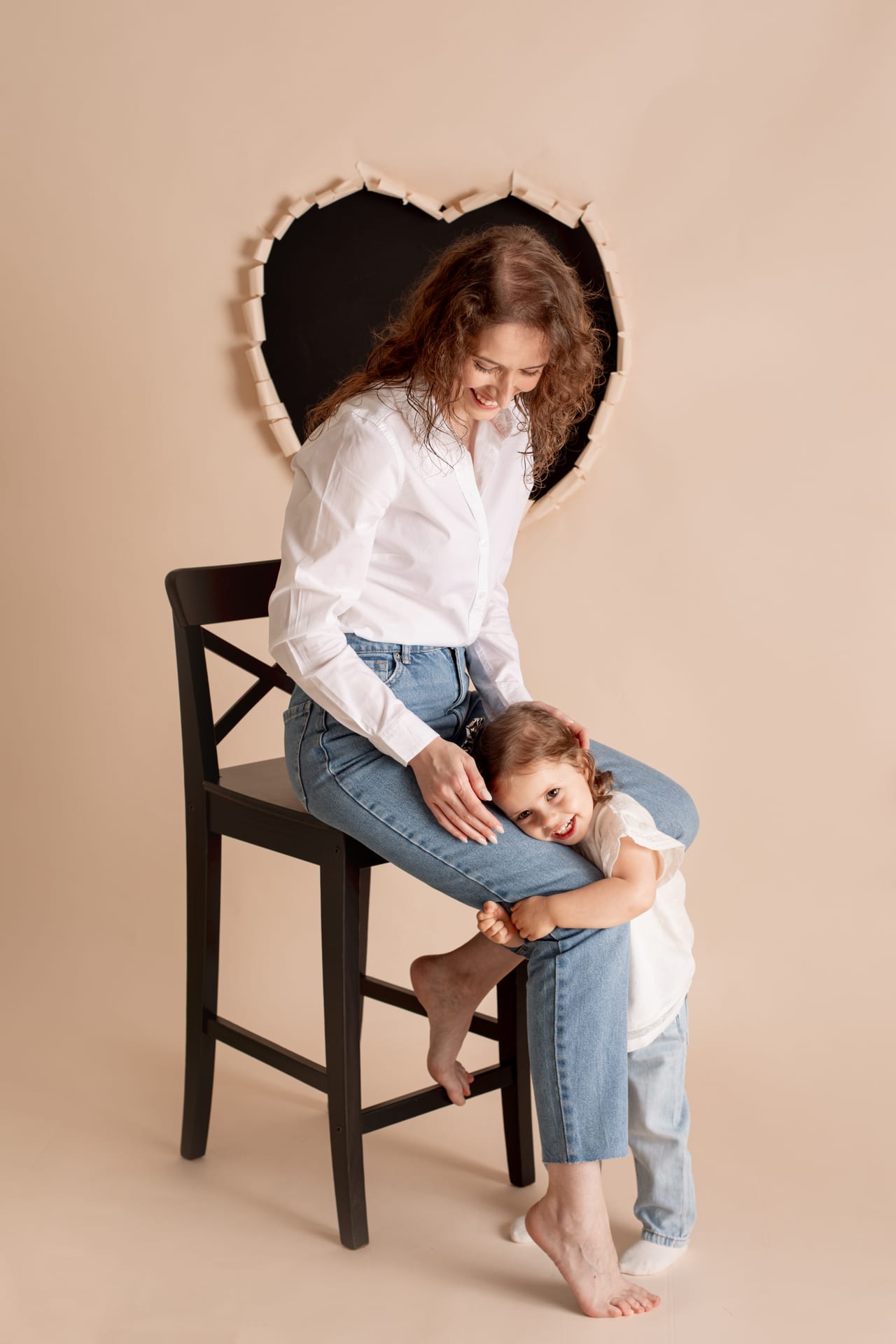 Mãe com camisa branca e jeans em banco preto alto com filha pequena agarrada à perna, fundo pêssego com decoração em forma de coração em estúdio, Susana Schmitz Fotografia