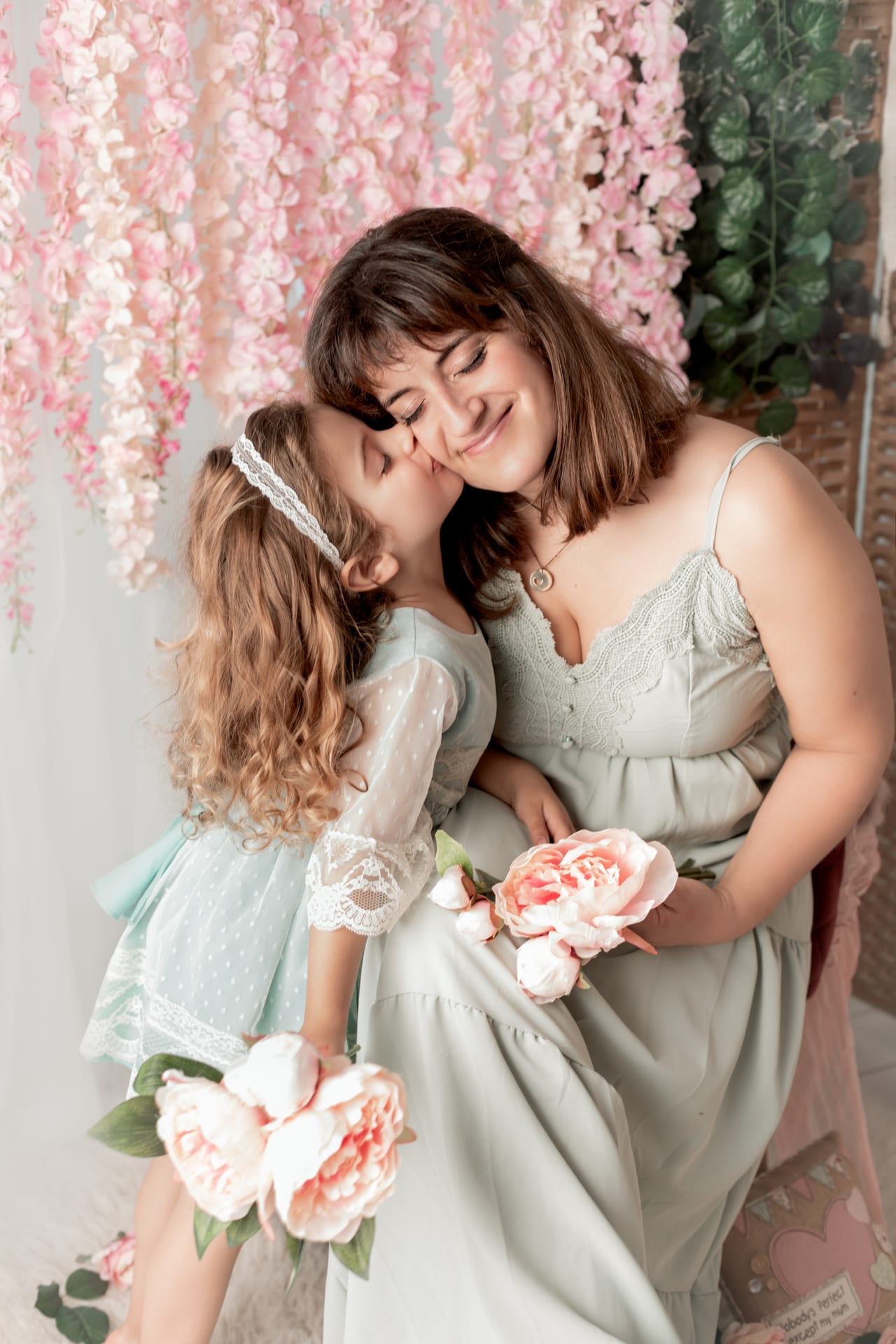 Filha pequena com vestido verde-água a beijar a mãe morena com vestido verde-sálvia, ambas com bouquet de flores cor-de-rosa, cenário de glicínias em estúdio, Susana Schmitz Fotografia, Porto