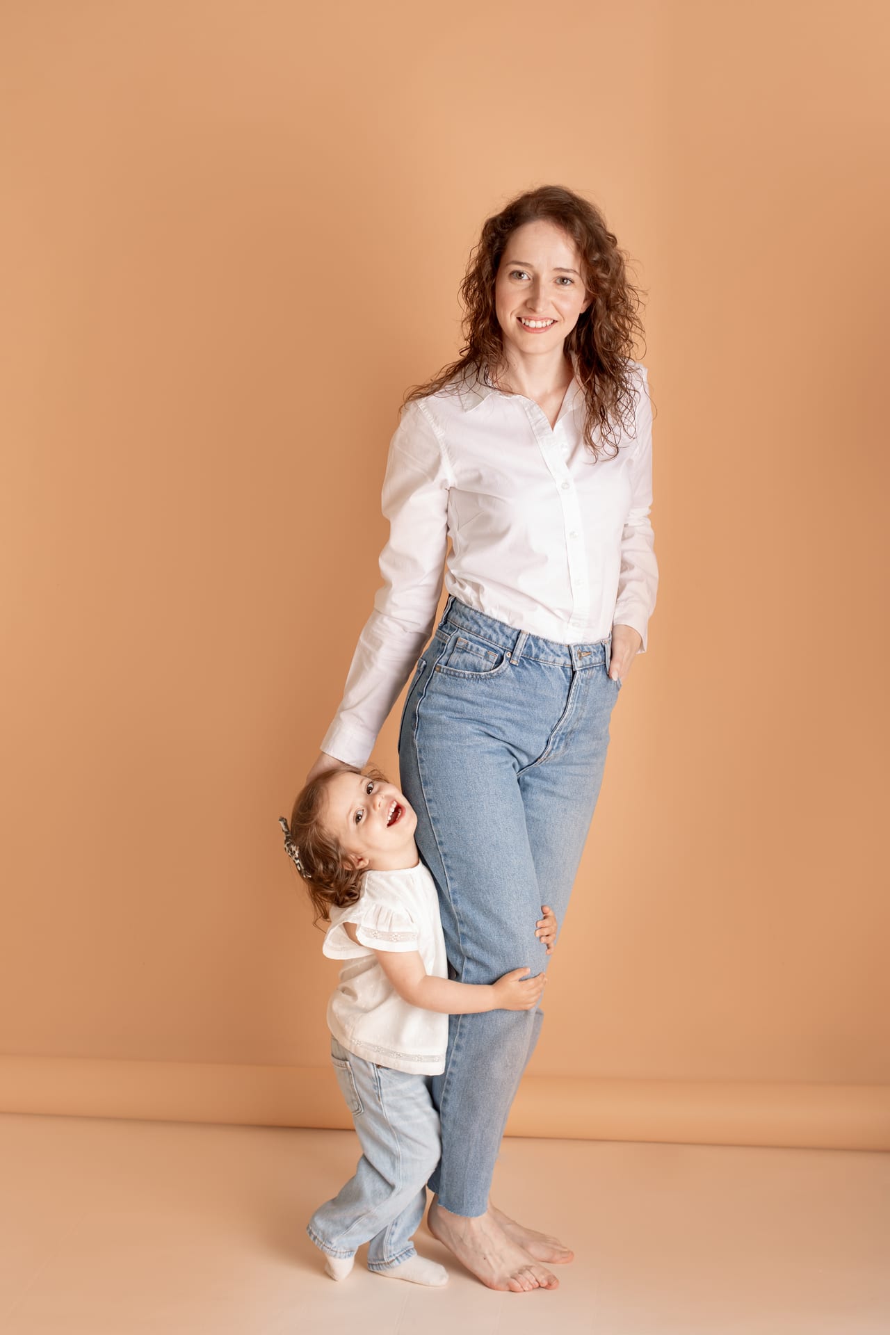 Mãe sorridente com camisa branca e jeans descalça com filha pequena agarrada à perna, fundo pêssego em estúdio, Susana Schmitz Fotografia, Porto