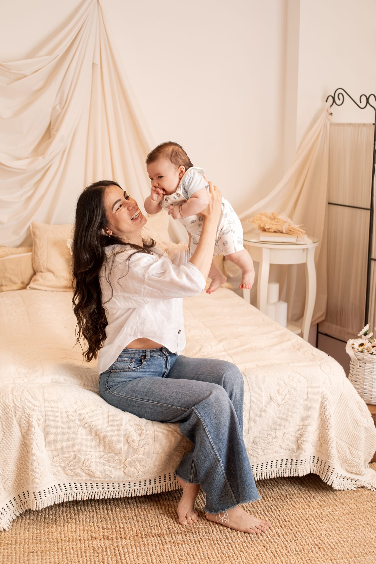 Mãe com camisa branca e jeans a levantar bebé com macacão branco em quarto com cama drapeada e cestos de vime, sessão de família em estúdio, Susana Schmitz Fotografia