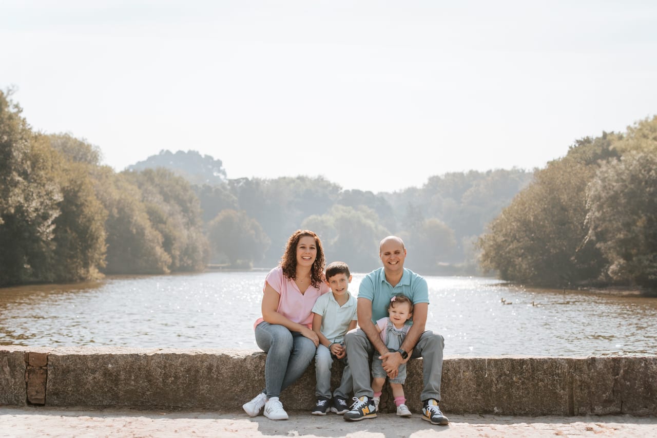 Família de quatro sentada em muro de pedra junto a lago num parque, pais e dois filhos em tons rosa e menta, sessão ao ar livre, Susana Schmitz Fotografia