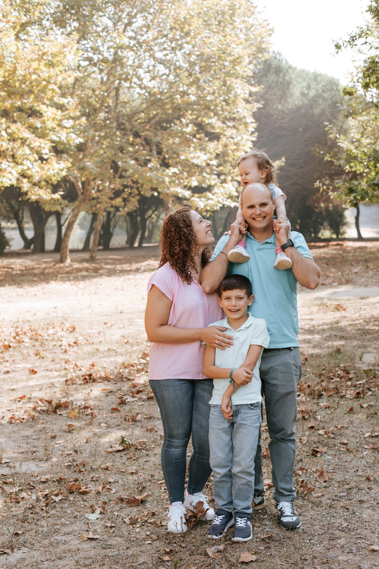 Família de quatro num parque ao ar livre no outono, pai com filha pequena aos ombros, mãe com top rosa e filho mais velho à frente, Susana Schmitz Fotografia, Porto