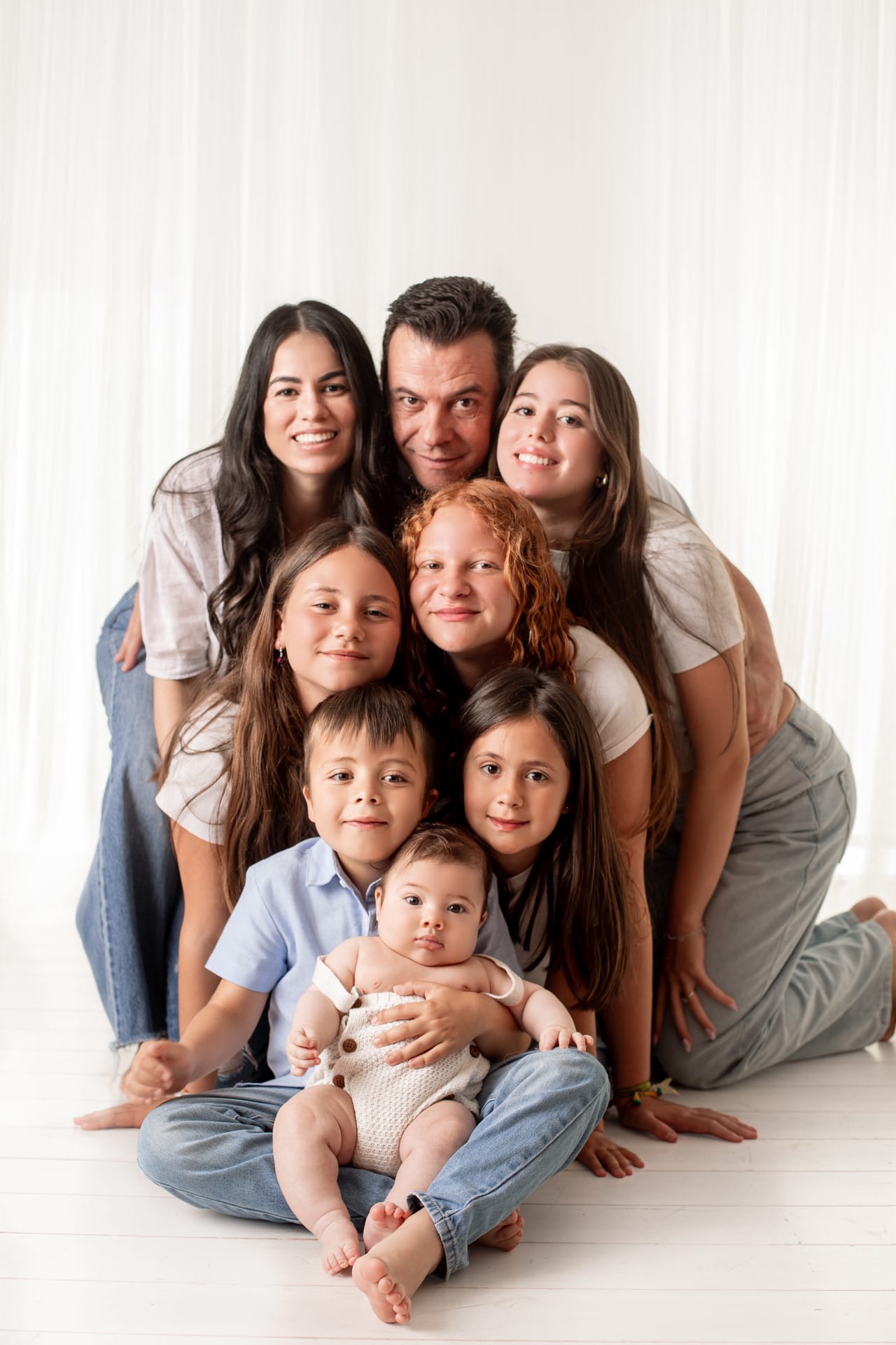 Família numerosa abraçada em estúdio com pais, três jovens, duas crianças e bebé ao colo, em tons neutros sobre fundo branco com cortinas, sessão de família, Susana Schmitz Fotografia, Porto