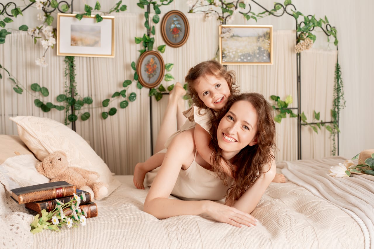 Mãe encaracolada e filha pequena deitadas numa cama de ferro forjado, fundo de quadros emoldurados e plantas trepadeiras, retrato íntimo em estúdio, Susana Schmitz Fotografia