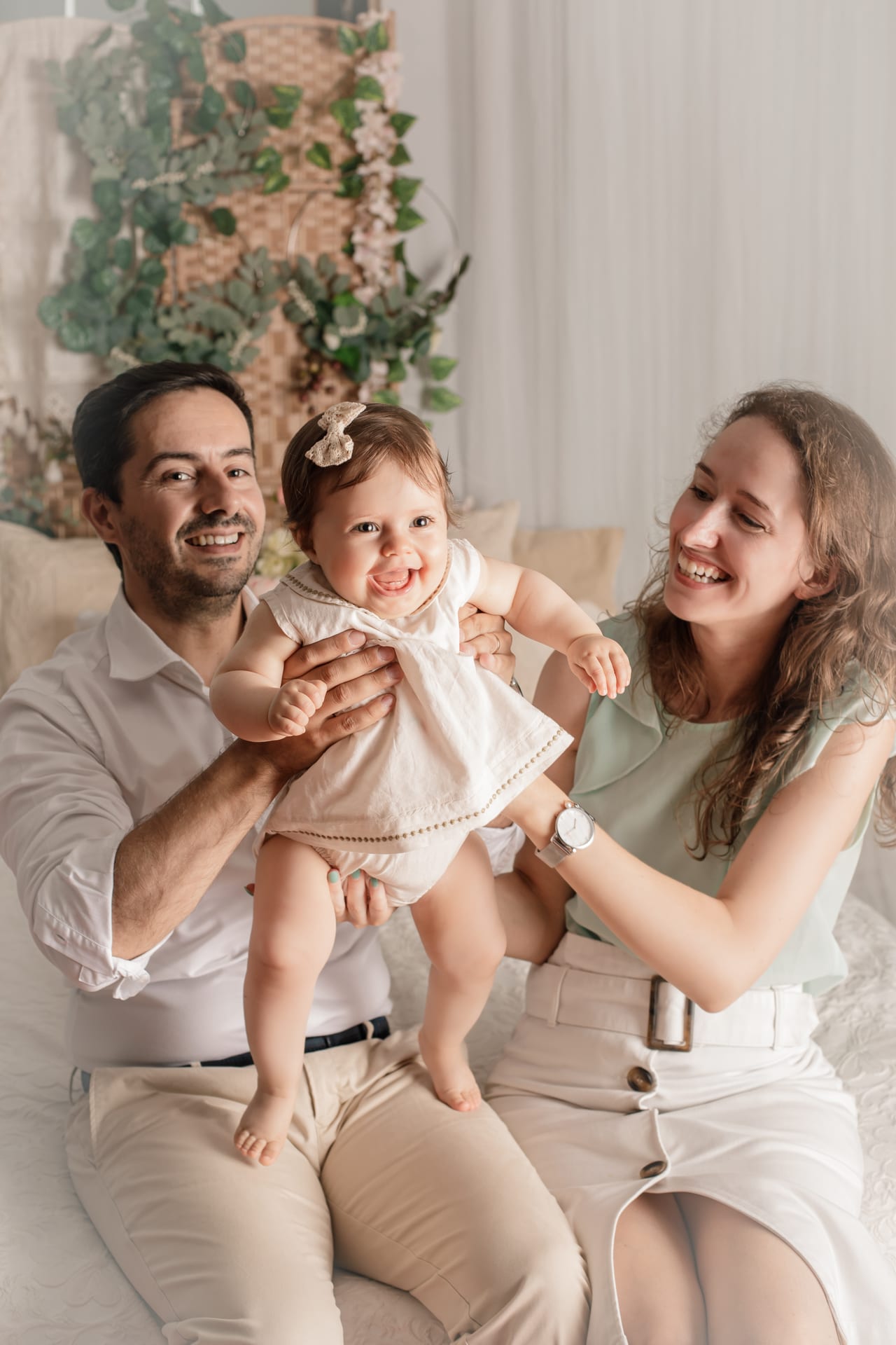 Pais sorridentes a segurar bebé com tiara num quarto em tons cremes com vegetação e cestos de vime, sessão de família em estúdio, Susana Schmitz Fotografia, Porto
