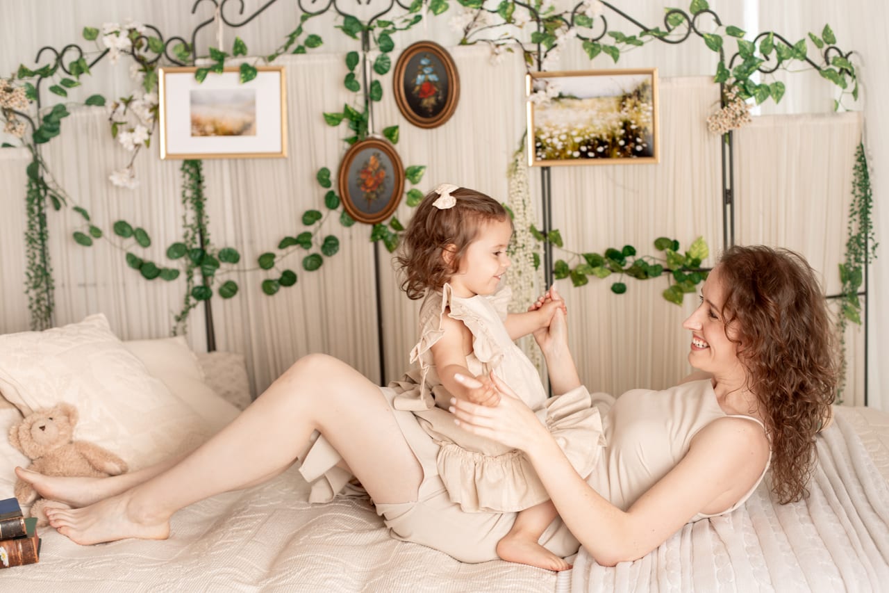 Mãe deitada com filha pequena ao colo numa cama de ferro forjado decorada com plantas trepadeiras e quadros emoldurados, cenário primaveril em estúdio, Susana Schmitz Fotografia