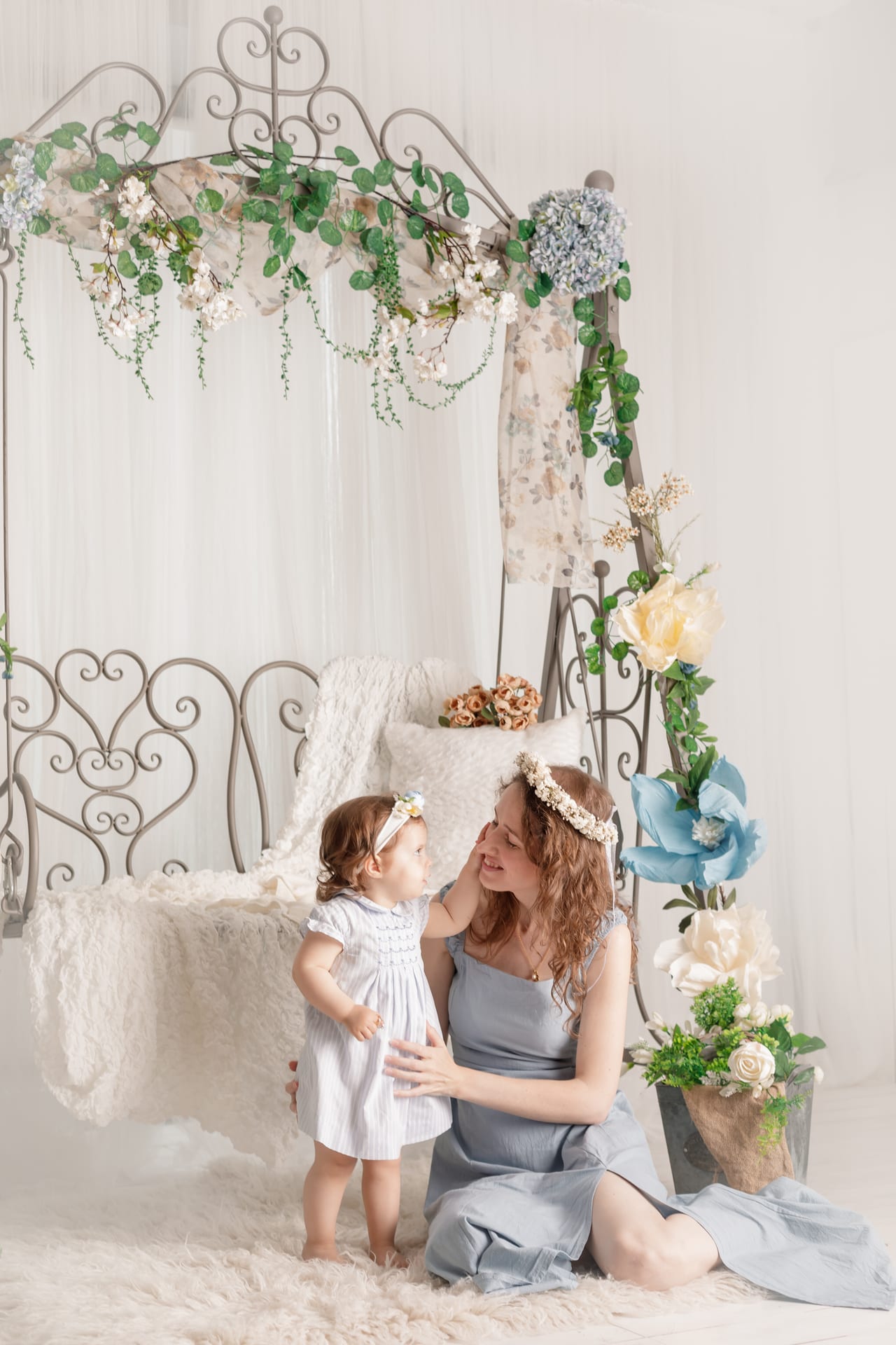 Mãe sentada com filha pequena em vestidos azul-cinza com coroa floral, cama de ferro forjado decorada com flores e folhagem, cenário primaveril em estúdio, Susana Schmitz Fotografia, Porto