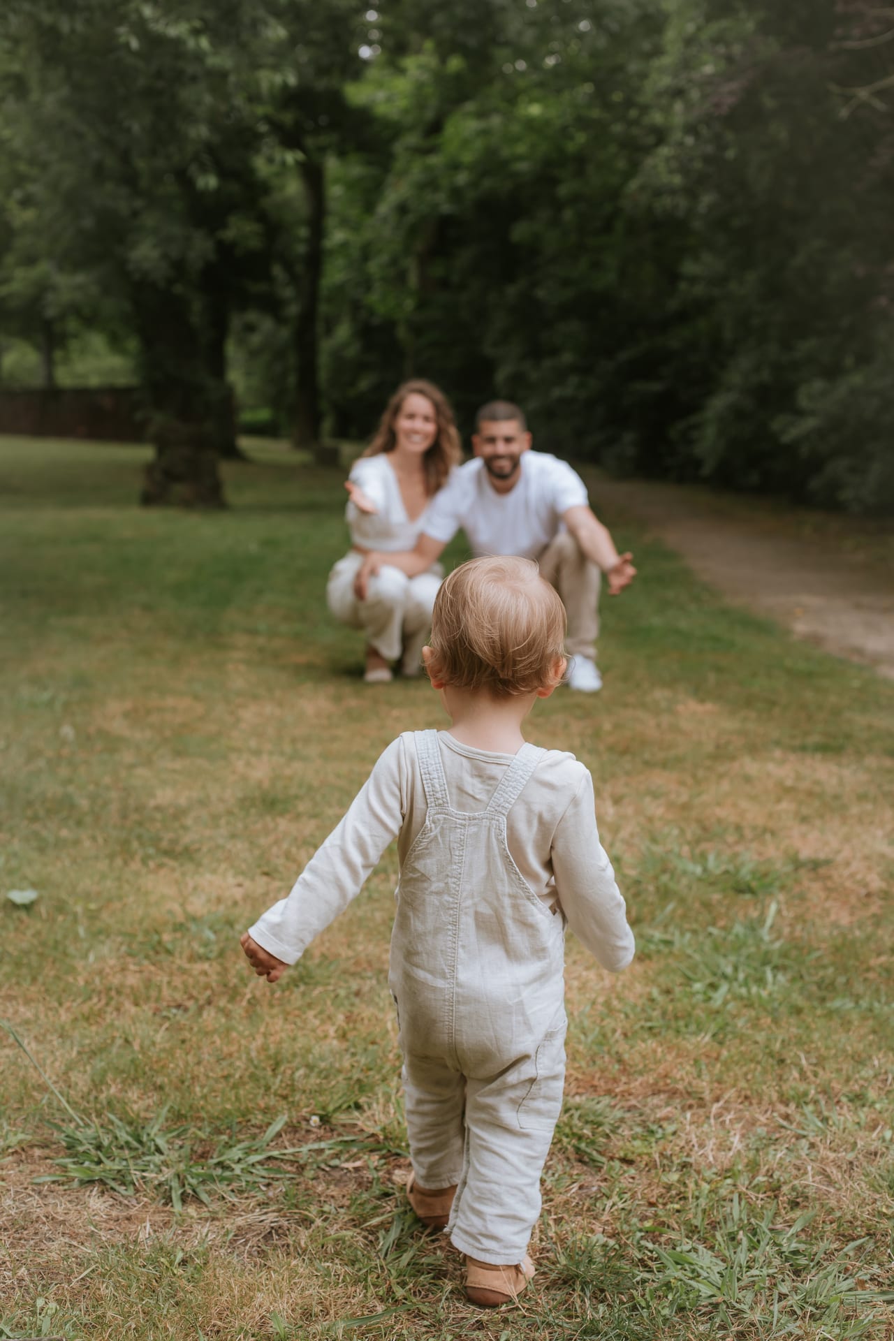 Bebé com macacão bege visto de costas a caminhar para os pais agachados num jardim verde, sessão de família ao ar livre, Susana Schmitz Fotografia, Porto