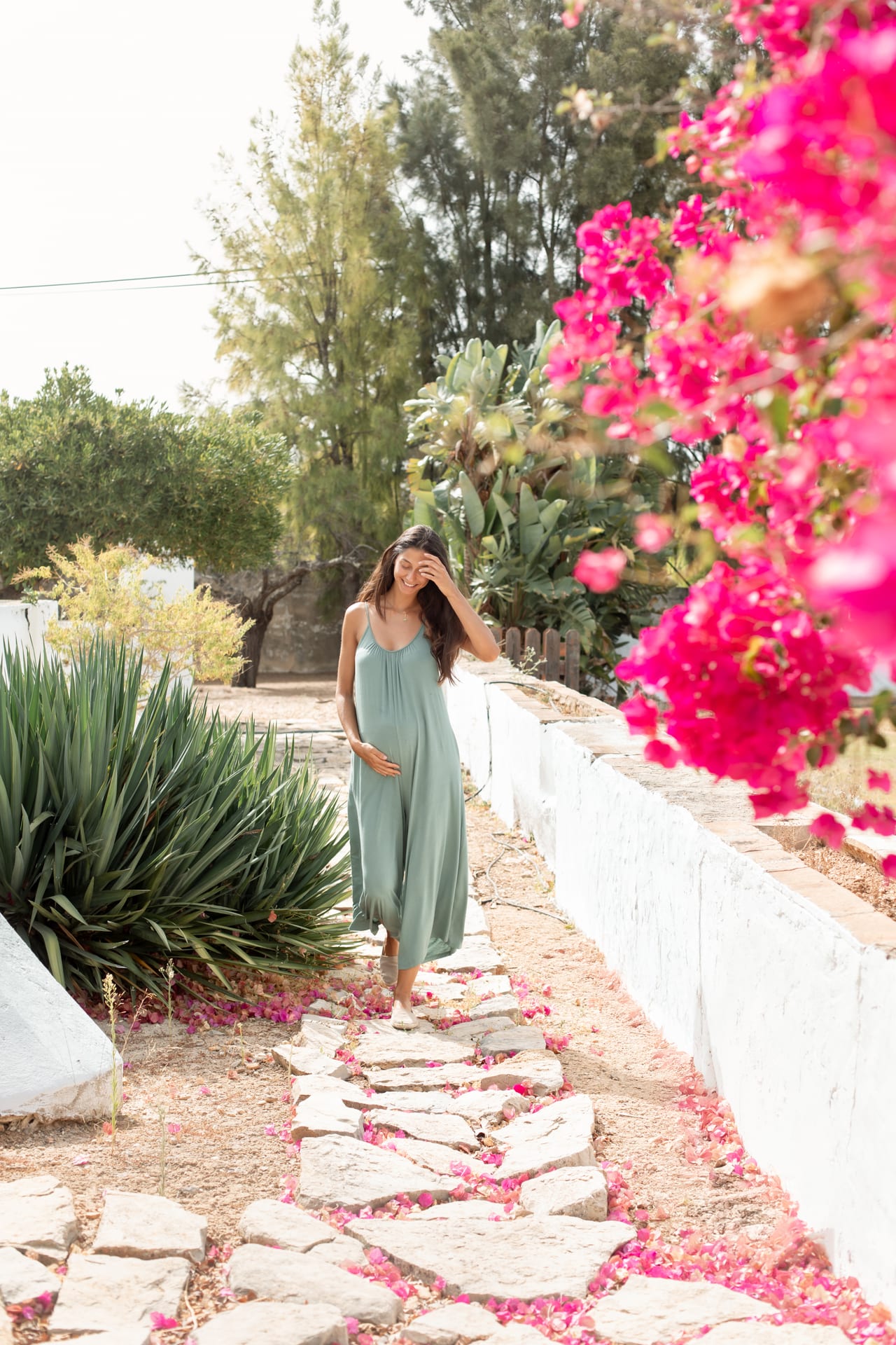 Grávida com macacão verde-sálvia em caminho de pedras com pétalas de buganvília rosa, sessão de gravidez ao ar livre em jardim, Susana Schmitz Fotografia, Porto