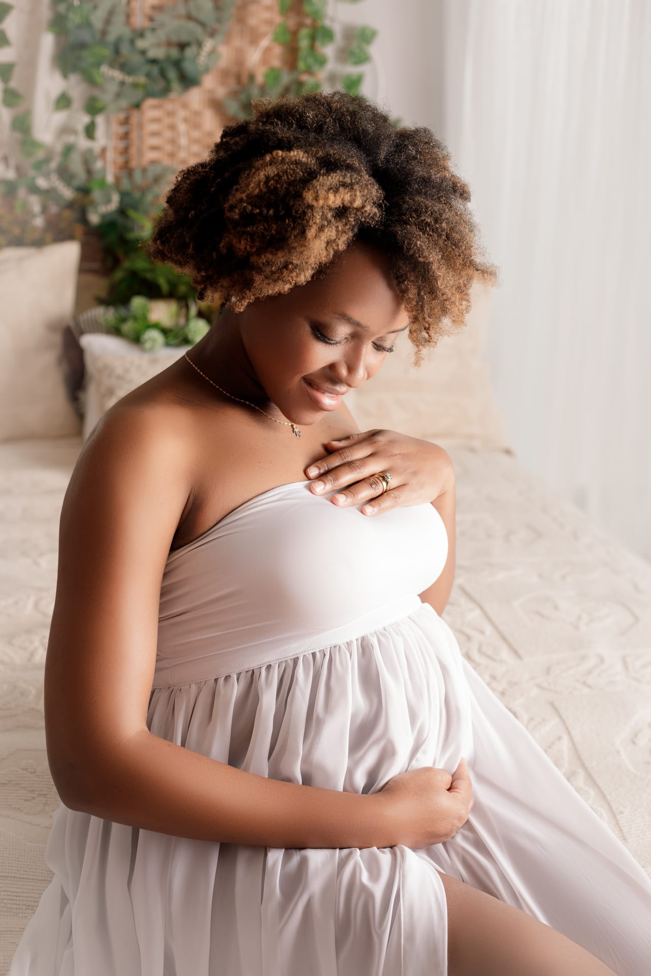 Grávida sentada com vestido branco drapeado, mãos sobre a barriga em estúdio luminoso com plantas, Susana Schmitz Fotografia, Porto