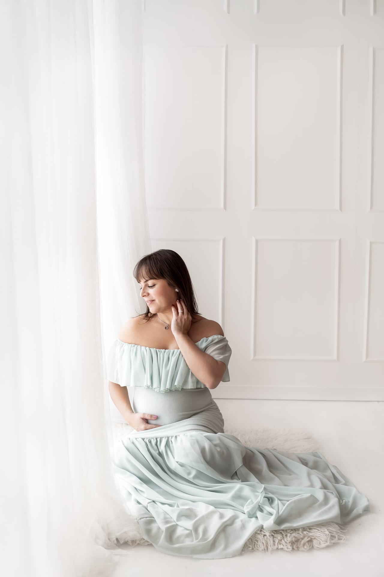 Grávida com vestido verde-água de ombros descobertos sentada no chão junto a cortinas brancas em estúdio luminoso, Susana Schmitz Fotografia, Porto