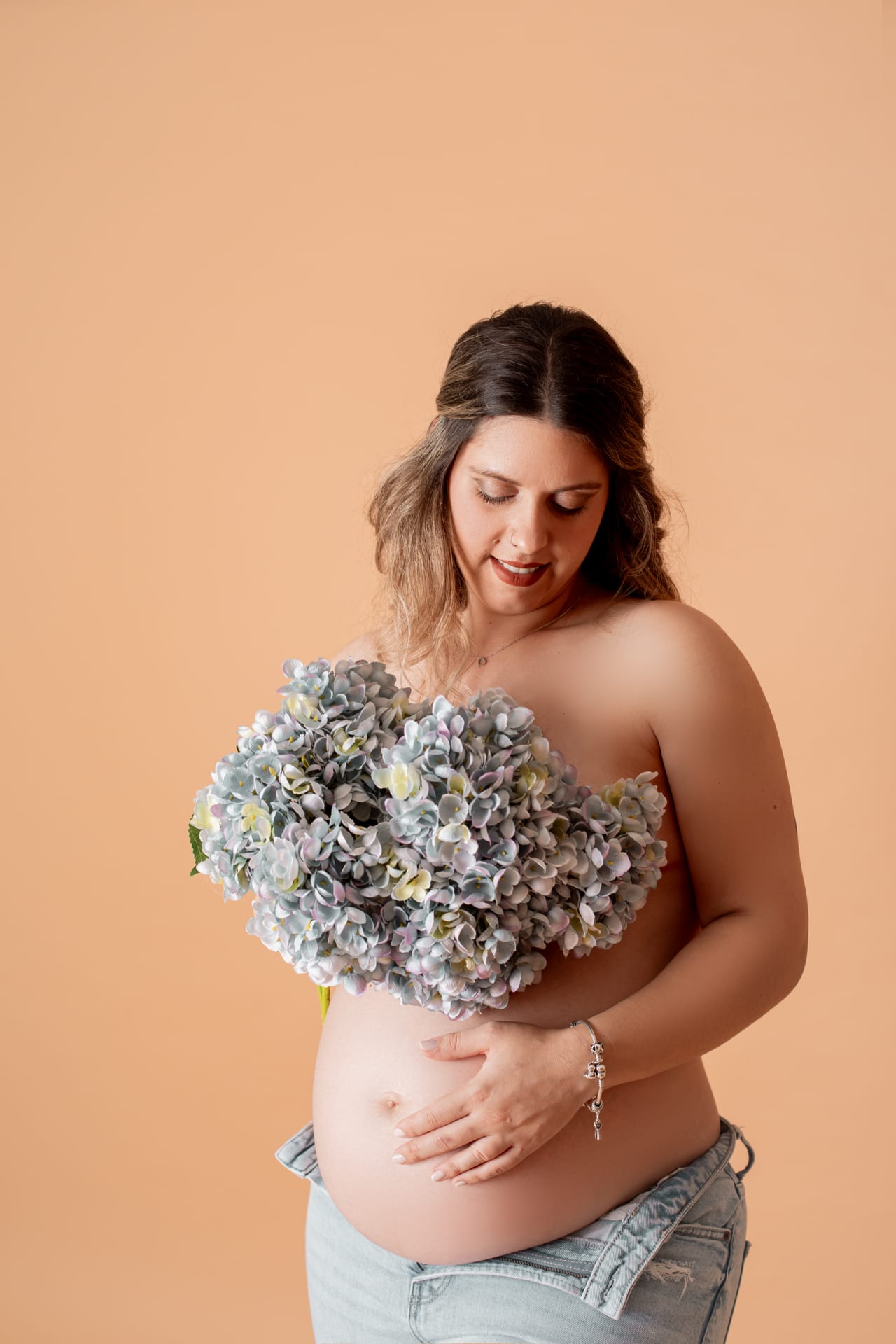 Grávida com cabelo solto a segurar ramo de hortênsias azul-pastel sobre o peito, em jeans, fundo pêssego em estúdio, Susana Schmitz Fotografia, Porto