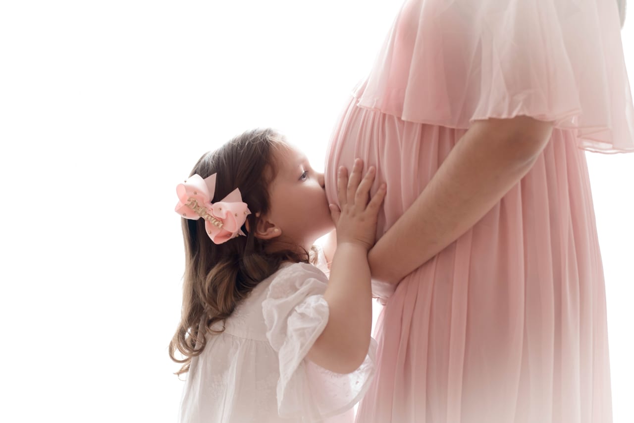 Filha com laço rosa a beijar a barriga da mãe grávida com vestido rosa pastel em estúdio de fundo branco, Susana Schmitz Fotografia