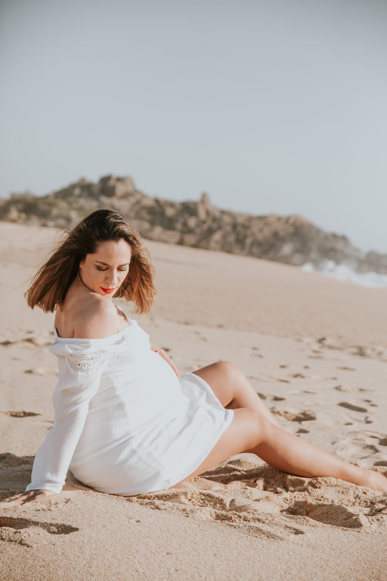Grávida em vestido branco sentada na areia da praia ao final da tarde, sessão de gravidez em natureza, Susana Schmitz Fotografia, Porto
