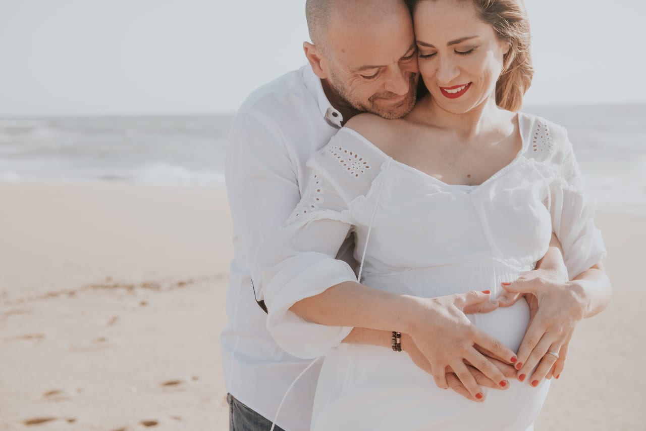 Casal grávido abraçado na praia, ambos com peças brancas, mãos sobrepostas na barriga, mar ao fundo, Susana Schmitz Fotografia