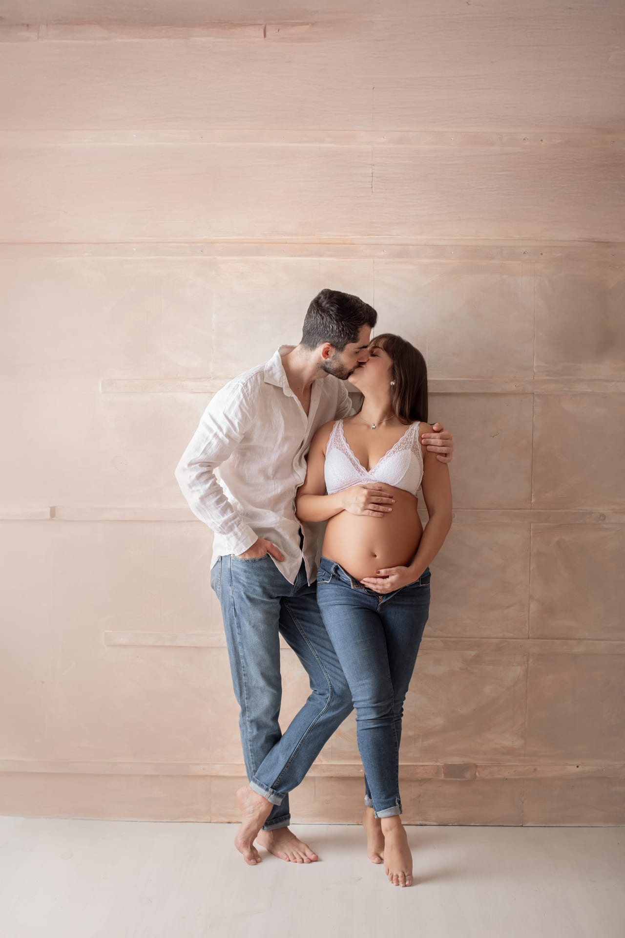 Casal a beijar-se em estúdio, ele de camisa branca e jeans, ela de soutien de renda branca e jeans com barriga à mostra, ambos descalços, Susana Schmitz Fotografia, Porto