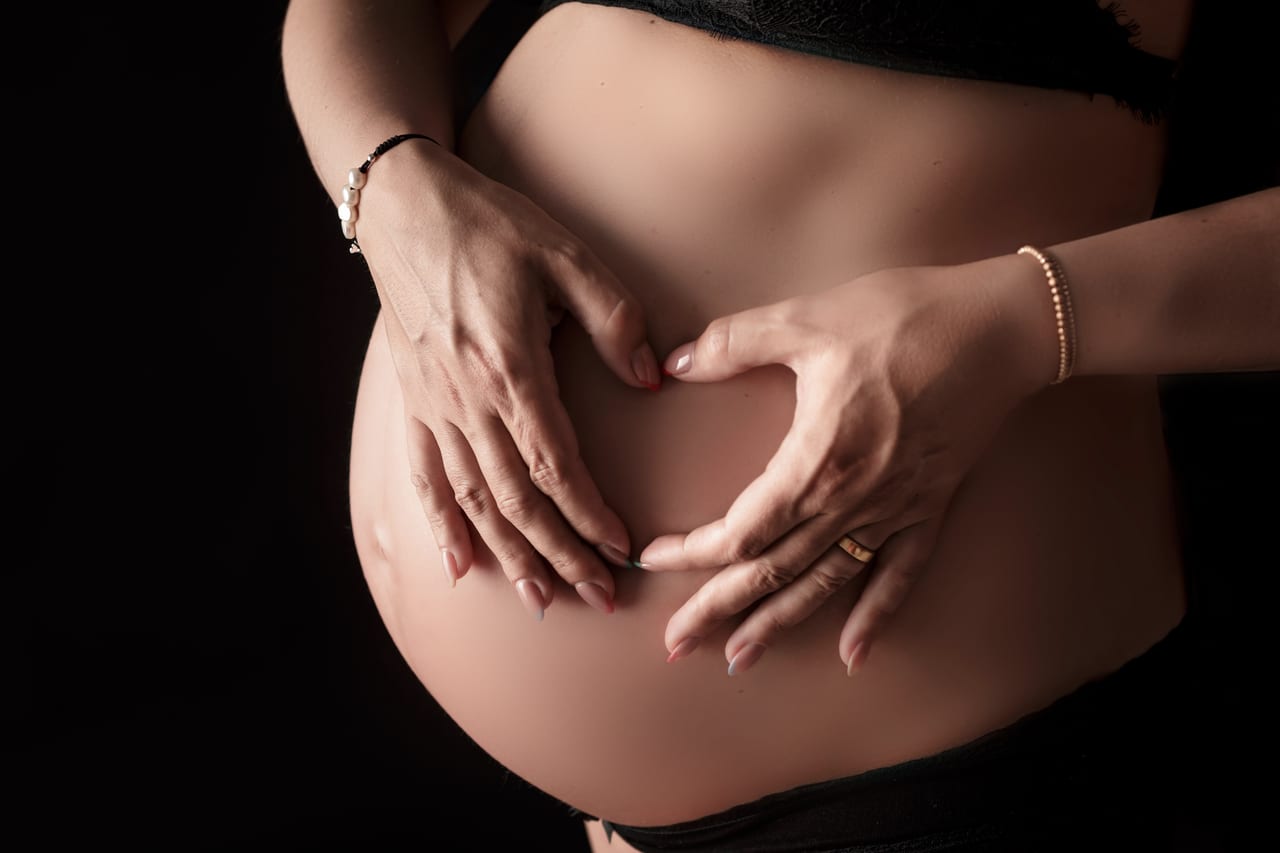 Detalhe das mãos dos pais a formarem um coração sobre a barriga de gravidez, fundo escuro em estúdio, Susana Schmitz Fotografia