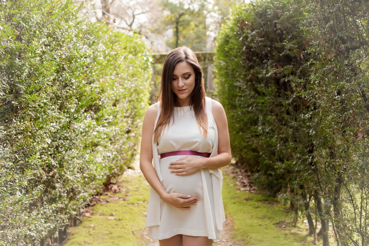 Grávida com vestido branco curto e cinto bordeaux num caminho de jardim entre sebes verdes, mãos na barriga, Susana Schmitz Fotografia