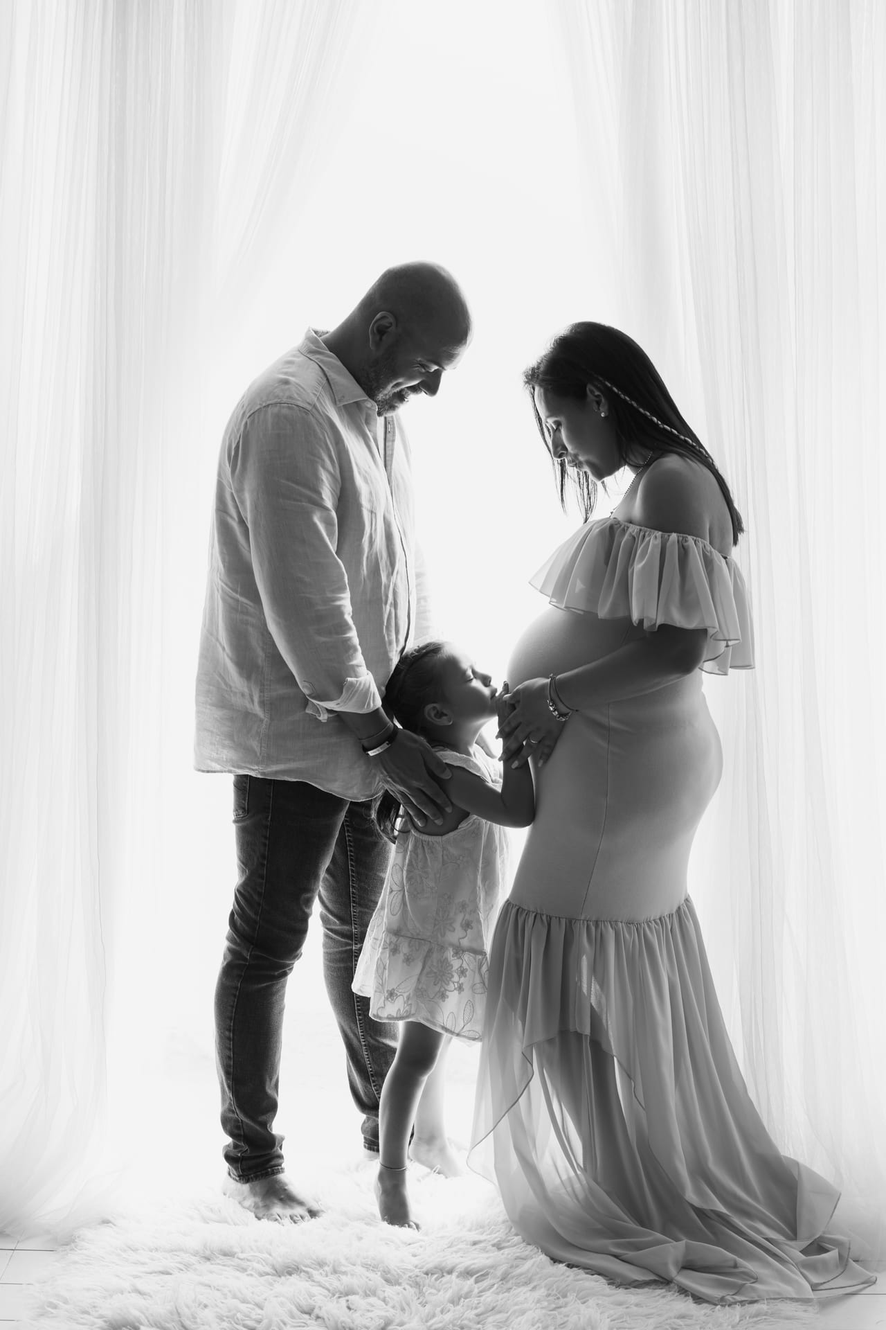 Família grávida em silhueta a contraluz junto a cortinas brancas, filha pequena a beijar a barriga da mãe, retrato a preto e branco em estúdio, Susana Schmitz Fotografia