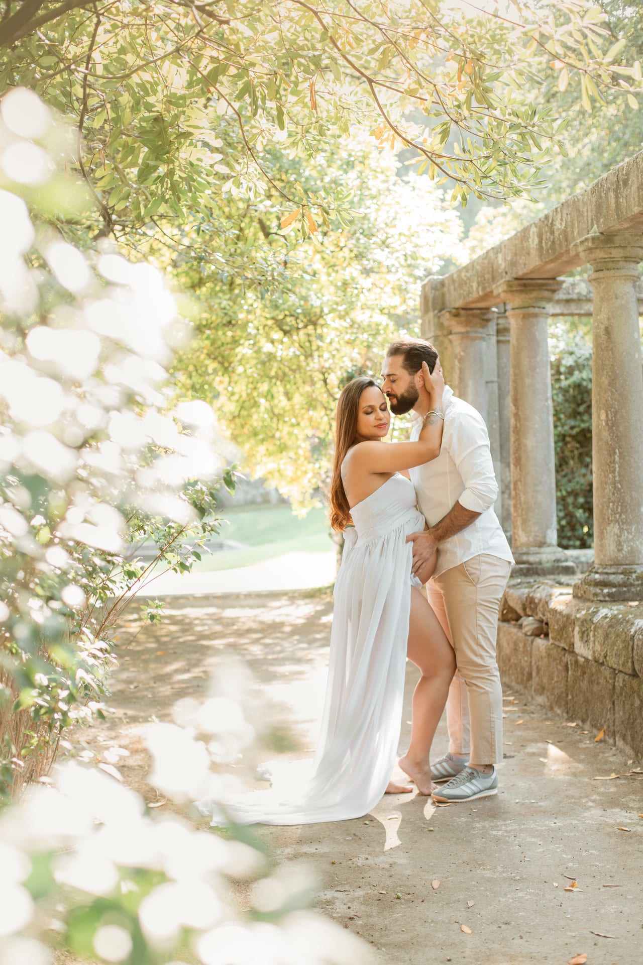Casal grávido abraçado entre pilares de pedra e folhagem na Quinta da Conceição, ela com vestido branco comprido, ele com camisa branca, Susana Schmitz Fotografia