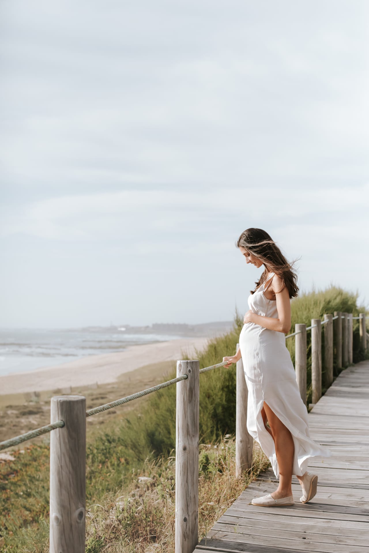 Grávida com vestido branco comprido encostada a guarda-corpo de madeira em passadiço junto a dunas e mar, vento no cabelo, Susana Schmitz Fotografia