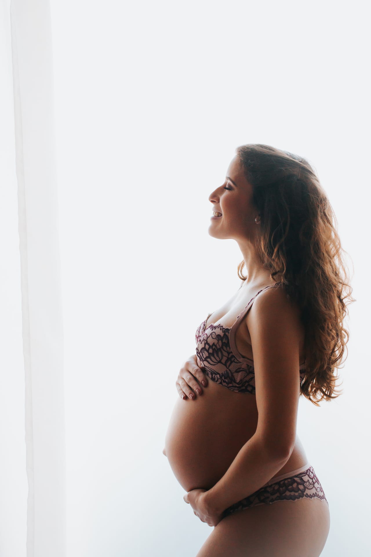 Grávida sorridente de perfil em contraluz junto a cortina branca, em lingerie de renda bordeaux, mãos a embalar a barriga, Susana Schmitz Fotografia, Porto
