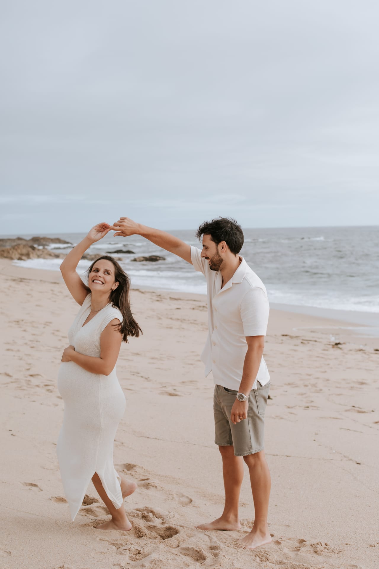 Casal grávido a dançar descalços na praia, ela com vestido branco comprido, ele a fazê-la rodopiar, mar ao fundo, Susana Schmitz Fotografia