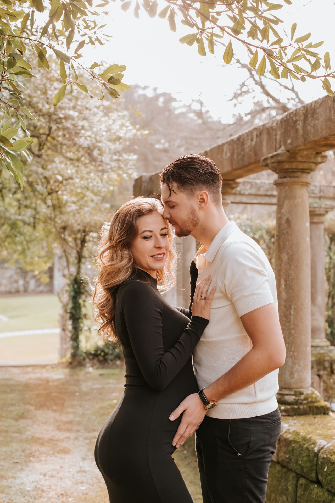 Casal grávido em jardim histórico com pilares de pedra, ele a beijar-lhe a testa, ela com vestido preto justo, sessão ao ar livre, Susana Schmitz Fotografia, Porto