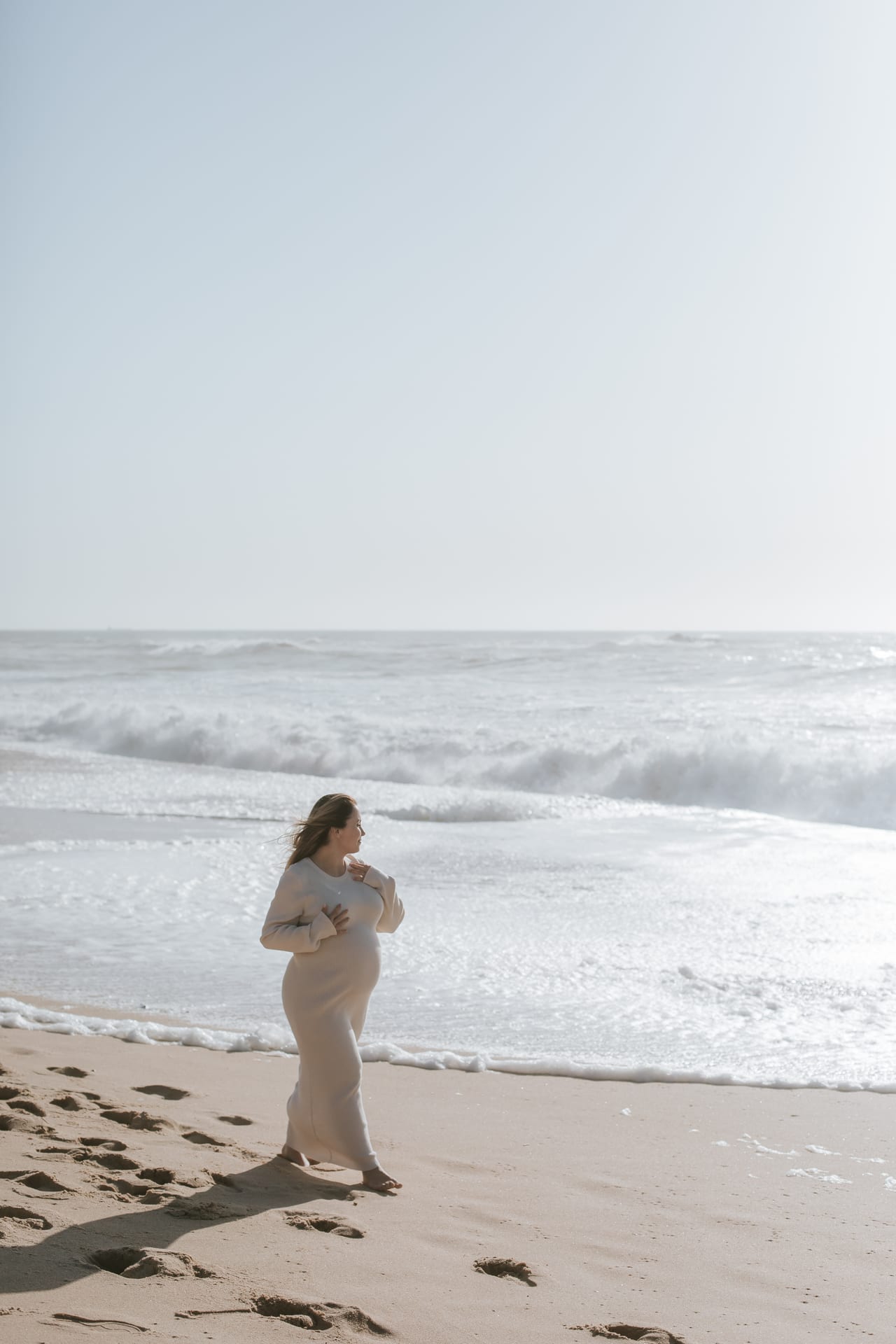 Grávida com vestido bege comprido a caminhar descalça na areia da praia, ondas atrás, sessão ao ar livre ao fim da tarde, Susana Schmitz Fotografia, Porto