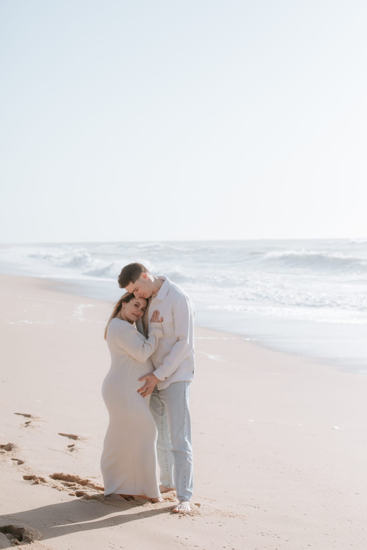 Casal grávido abraçado na praia ao fim da tarde, ela com vestido bege, ondas ao fundo, Susana Schmitz Fotografia, Porto