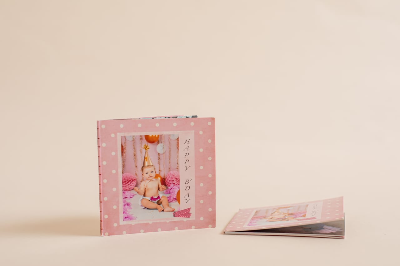 Mini-livro com capa rosa de bolinhas e título "Happy B.Day" com retrato de bebé em sessão de smash the cake, ao lado de outro mini-livro aberto, produto Susana Schmitz Fotografia, Porto