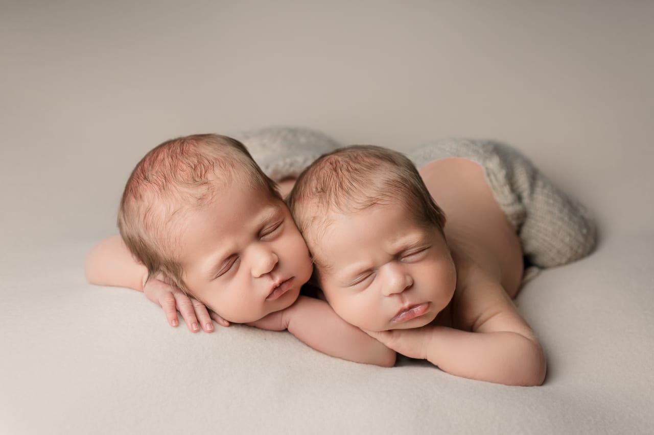 Gémeos recém-nascidos a dormir lado-a-lado com cabeças apoiadas nas mãos sobre fundo bege liso, sessão de gémeos em estúdio, Susana Schmitz Fotografia