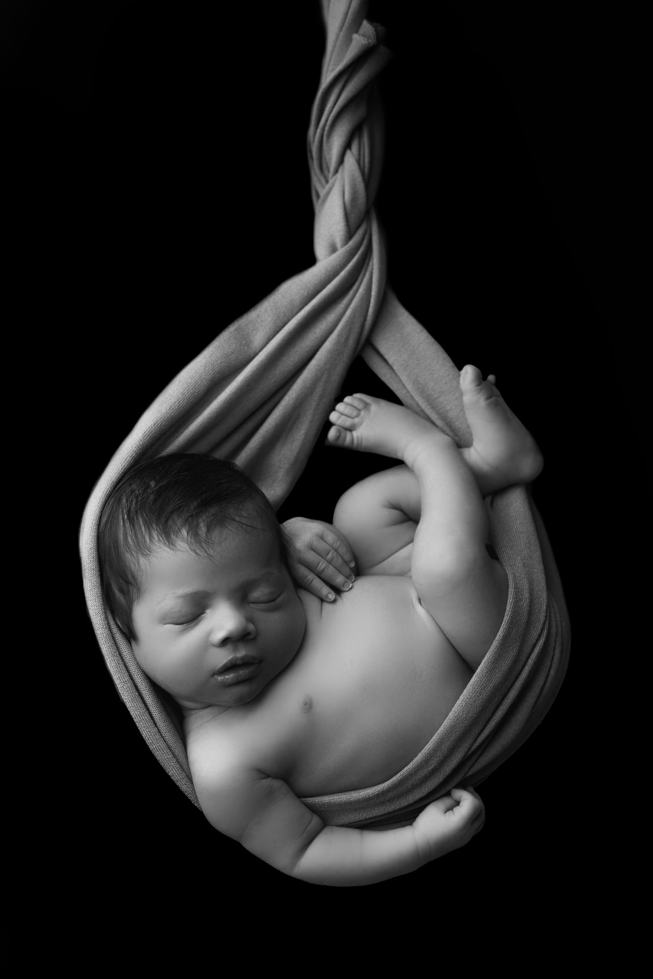 Recém-nascido suspenso em wrap pendurado em cocoon, retrato dramático a preto e branco contra fundo escuro, Susana Schmitz Fotografia