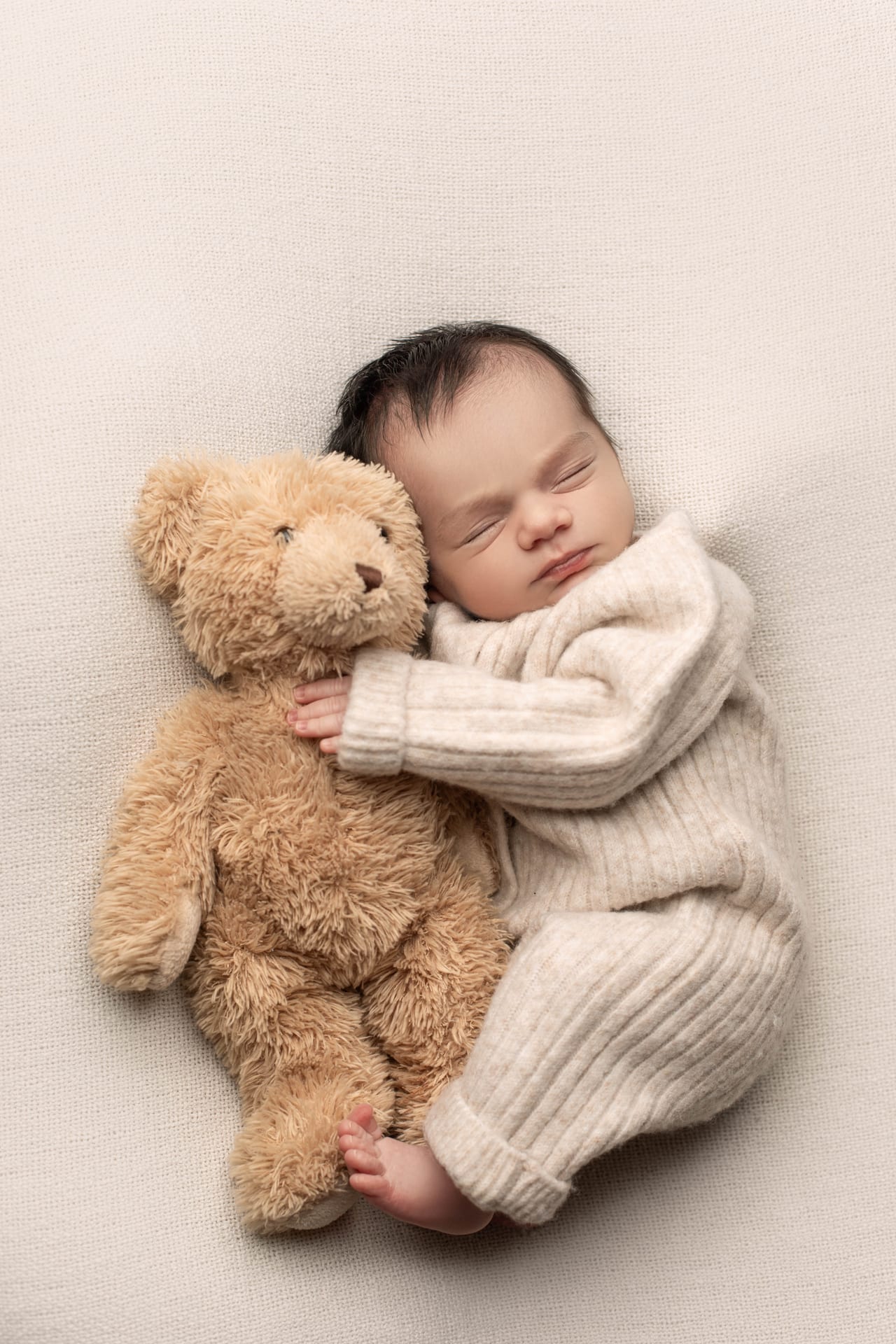 Bebé recém-nascido com fato de tricô bege a abraçar urso castanho de peluche em pose lateral sobre fundo bege liso, Susana Schmitz Fotografia