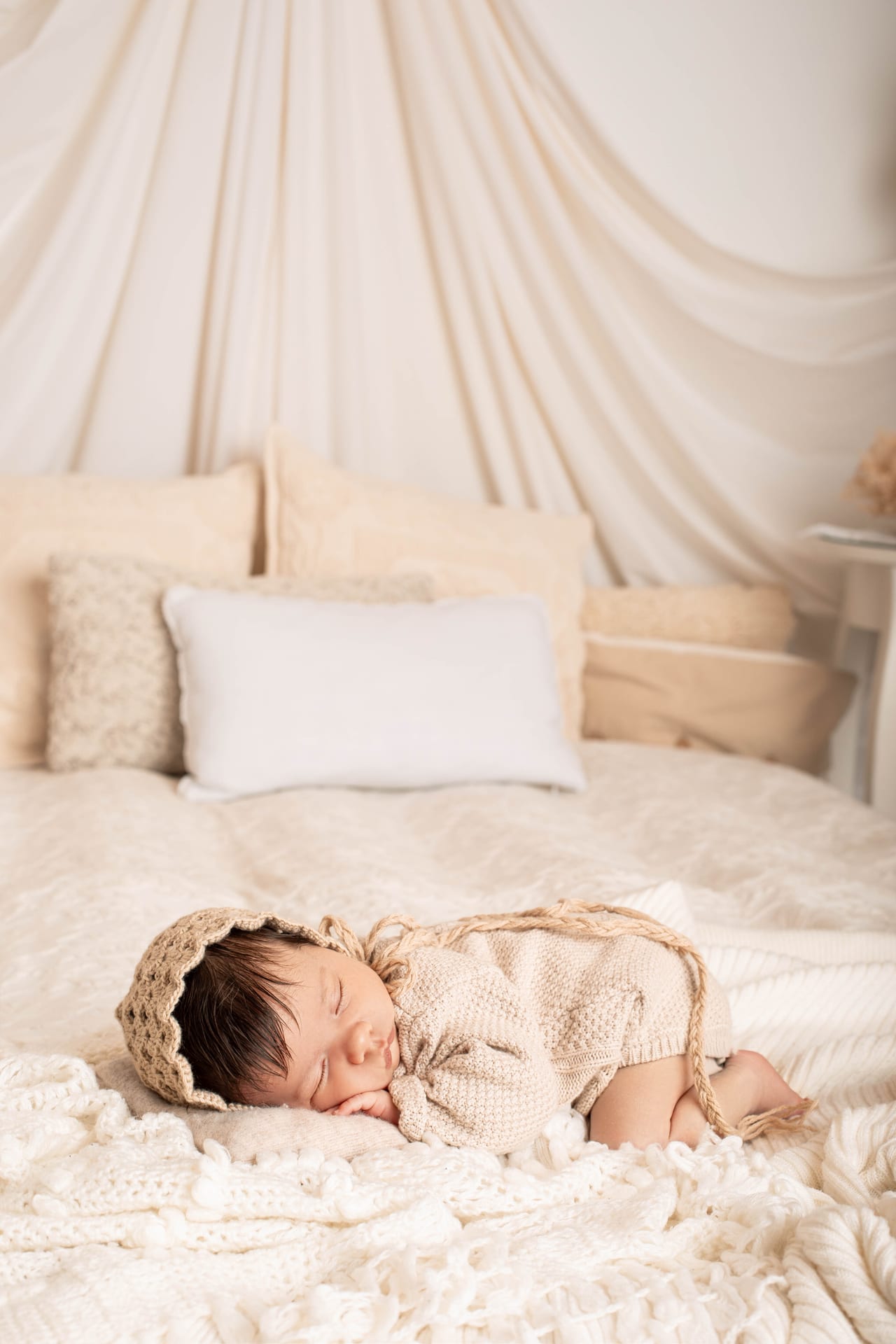 Bebé recém-nascido com touca de croché bege e fato de tricô a dormir em cama com cortinas e almofadas creme em ambiente de quarto, Susana Schmitz Fotografia