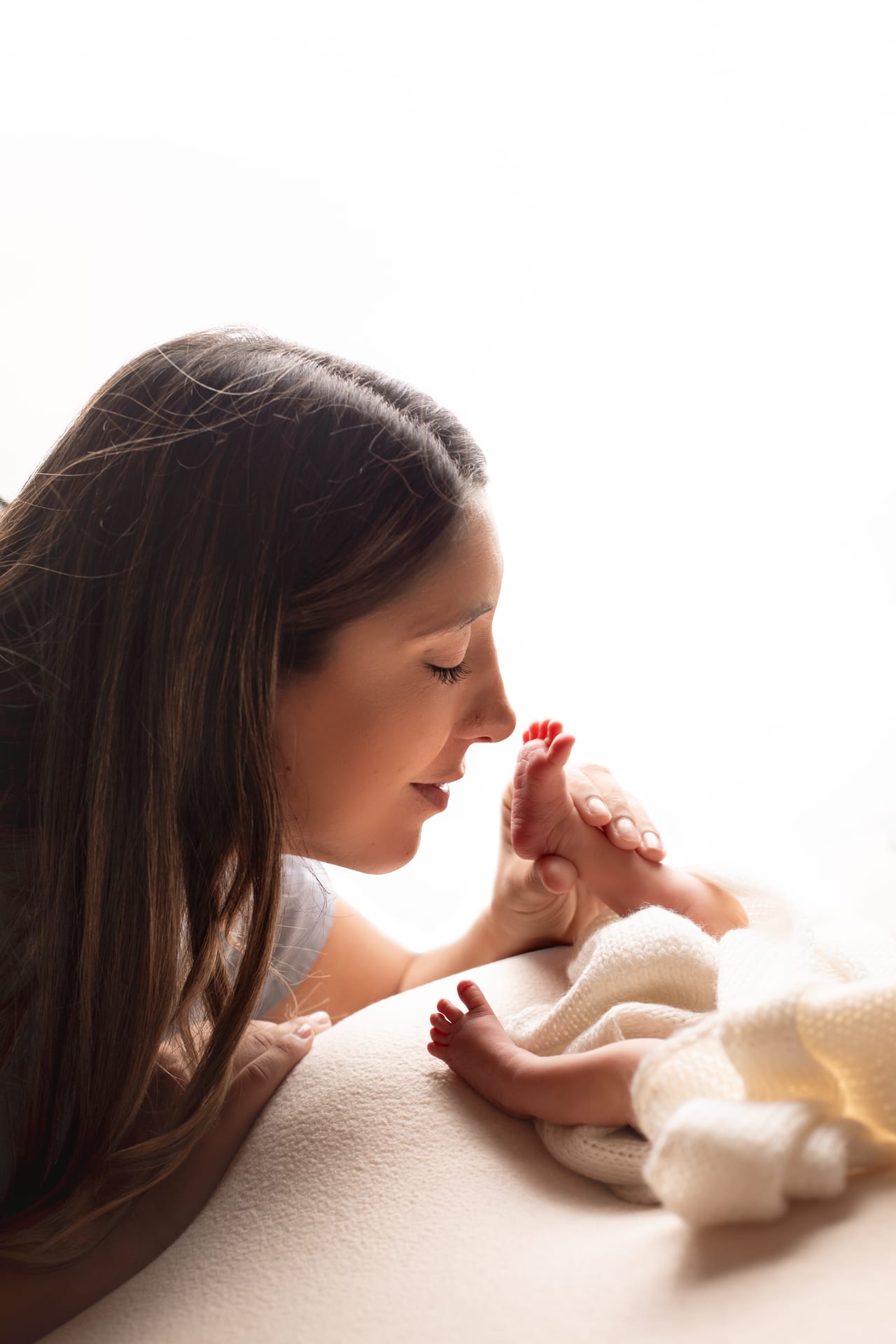 Mãe a beijar os pés do bebé recém-nascido envolto em manta branca, contraluz suave em estúdio, Susana Schmitz Fotografia, Porto